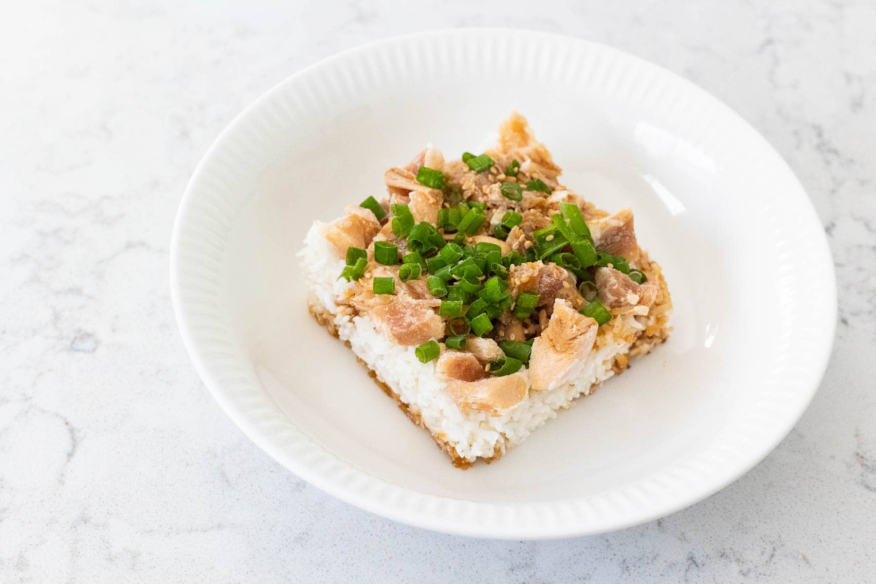 A serving of Rotisserie Chicken Teriyaki Rice Bowl with white rice topped with shredded chicken, teriyaki sauce, sesame seeds, and chopped green onions in a white bowl.