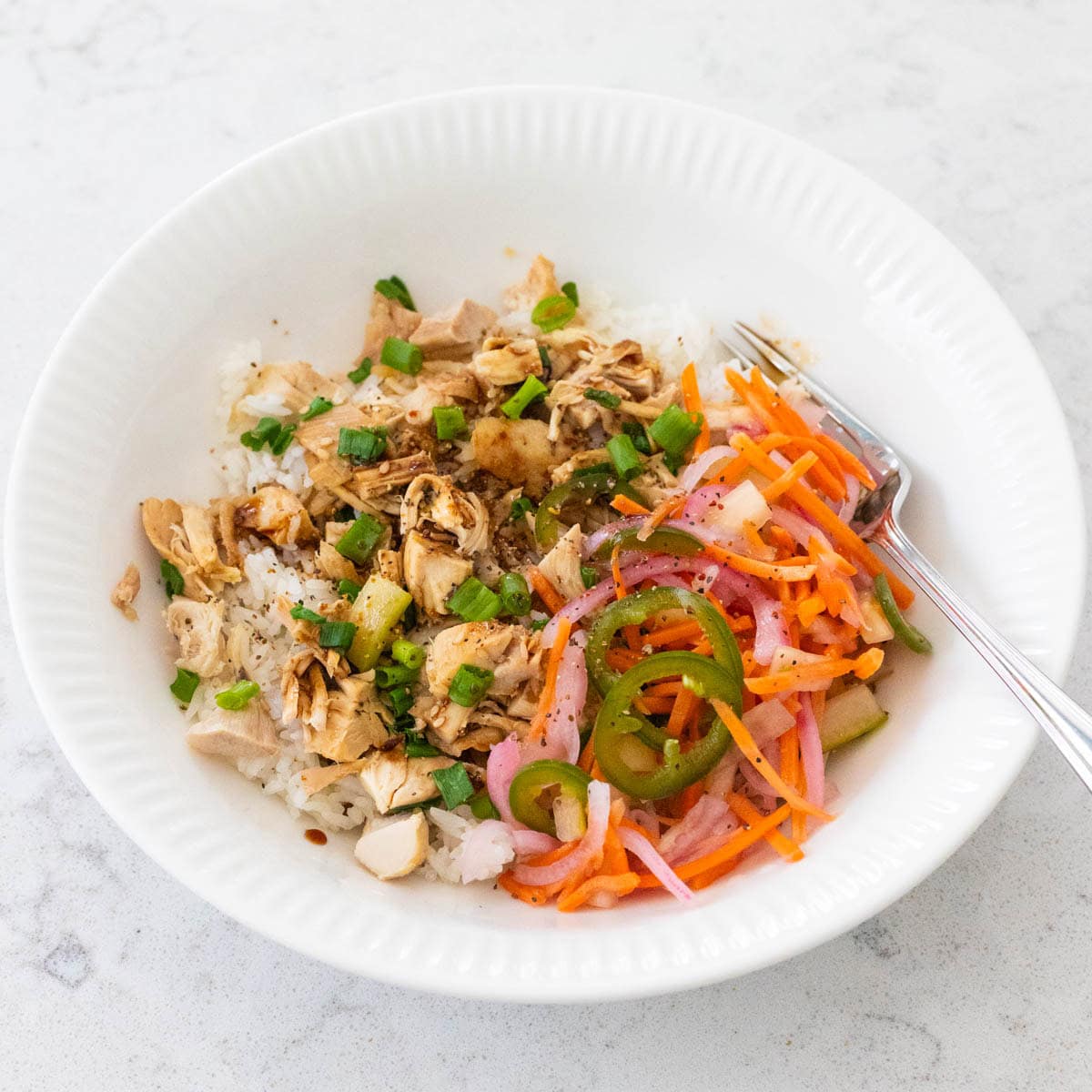 A white bowl filled with rice topped with shredded rotisserie chicken, teriyaki sauce, green onions, and a side of quick pickled carrots, red onion, and jalapeño slices.