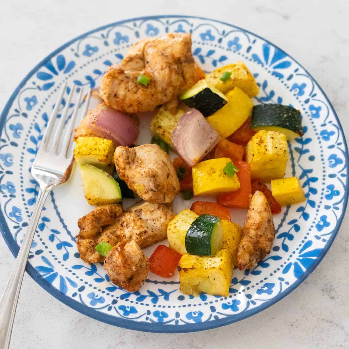 Plate of baked seasoned chicken pieces with roasted zucchini, yellow squash, red bell pepper, and red onion served on a blue patterned dish with a fork.