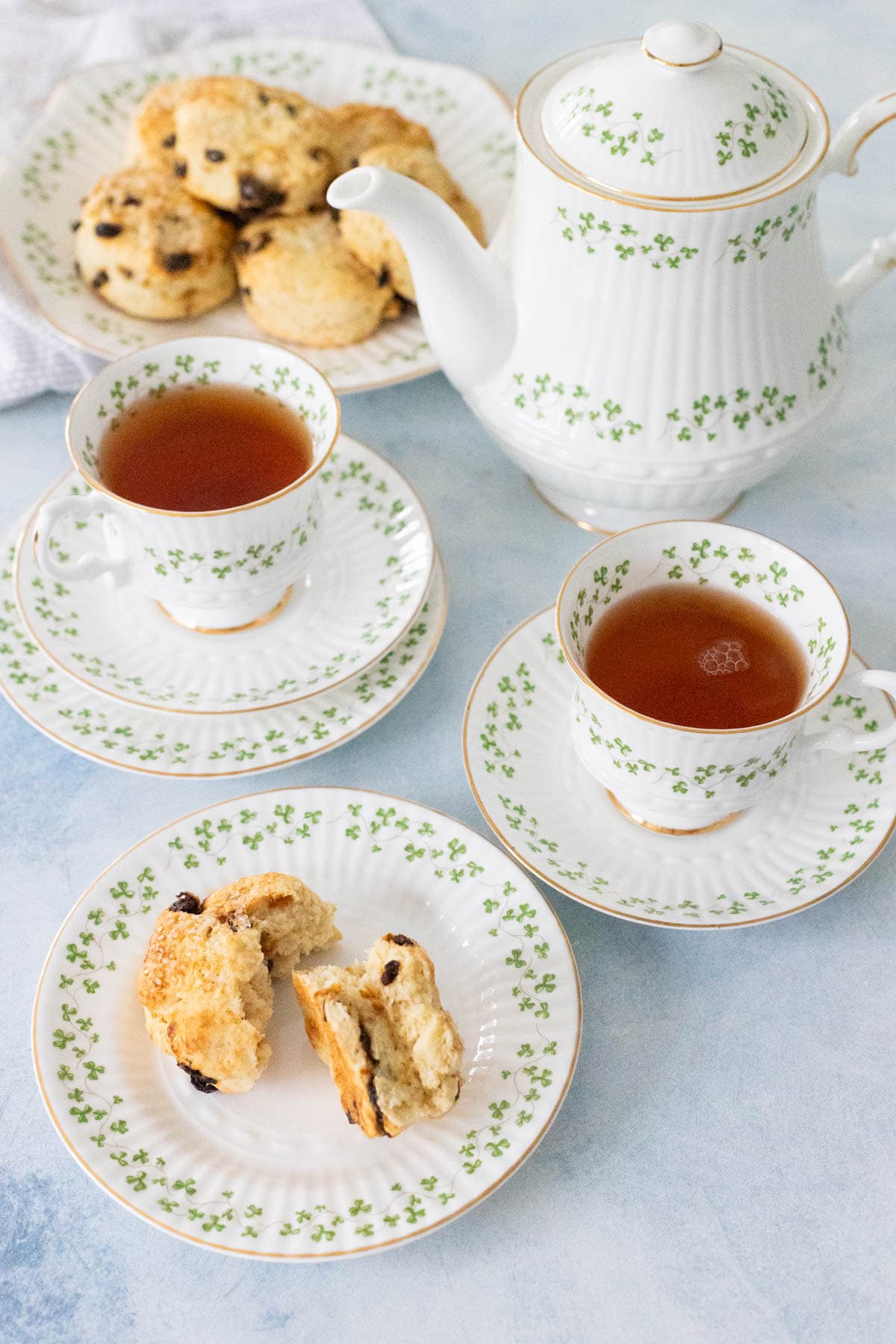 A tea pot and tea cups filled with tea have delicate shamrocks around the rims of the dishes. A plate of scones rests in the back.