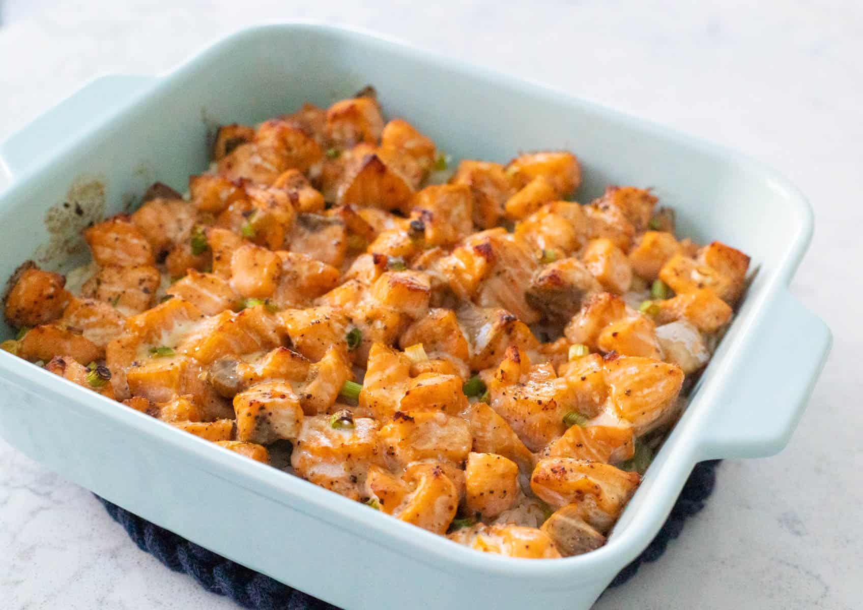 Spicy salmon sushi bake in a light blue baking dish with cubed salmon and green onions over seasoned rice.
