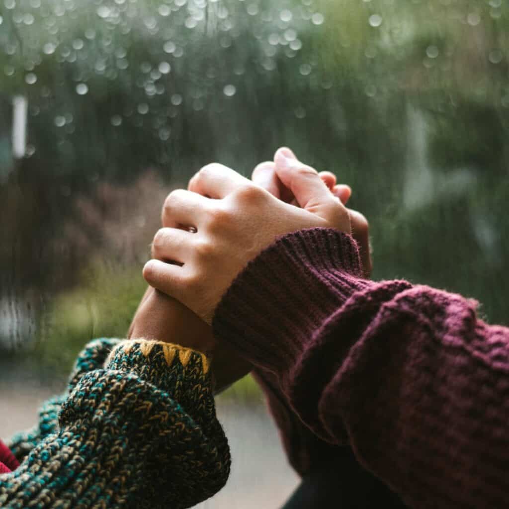 A close up of two people holding hands in front of a rainy window.
