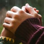 A close up of two people holding hands in front of a rainy window.