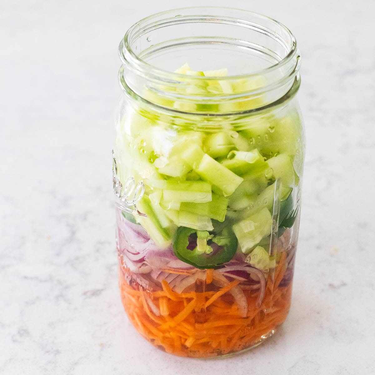 A mason jar filled with quick pickled vegetables, including sliced red onion, shredded carrots and jalapeño rounds submerged in brine.