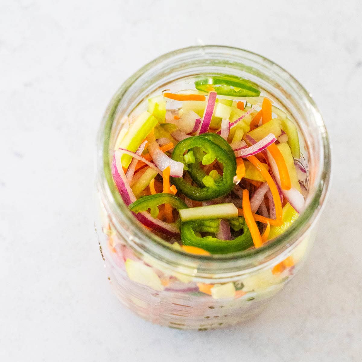A mason jar is filled with mixed pickled vegetables including shredded carrots, cucumbers, and jalapeno.