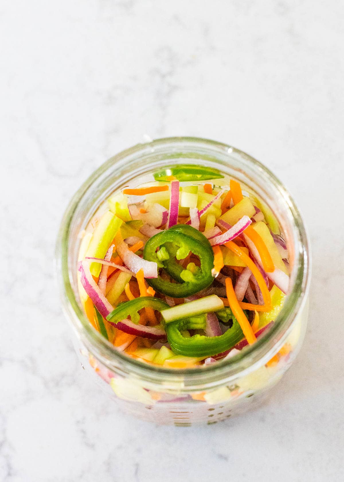 A mason jar is filled with mixed pickled vegetables including shredded carrots, cucumbers, and jalapeno.