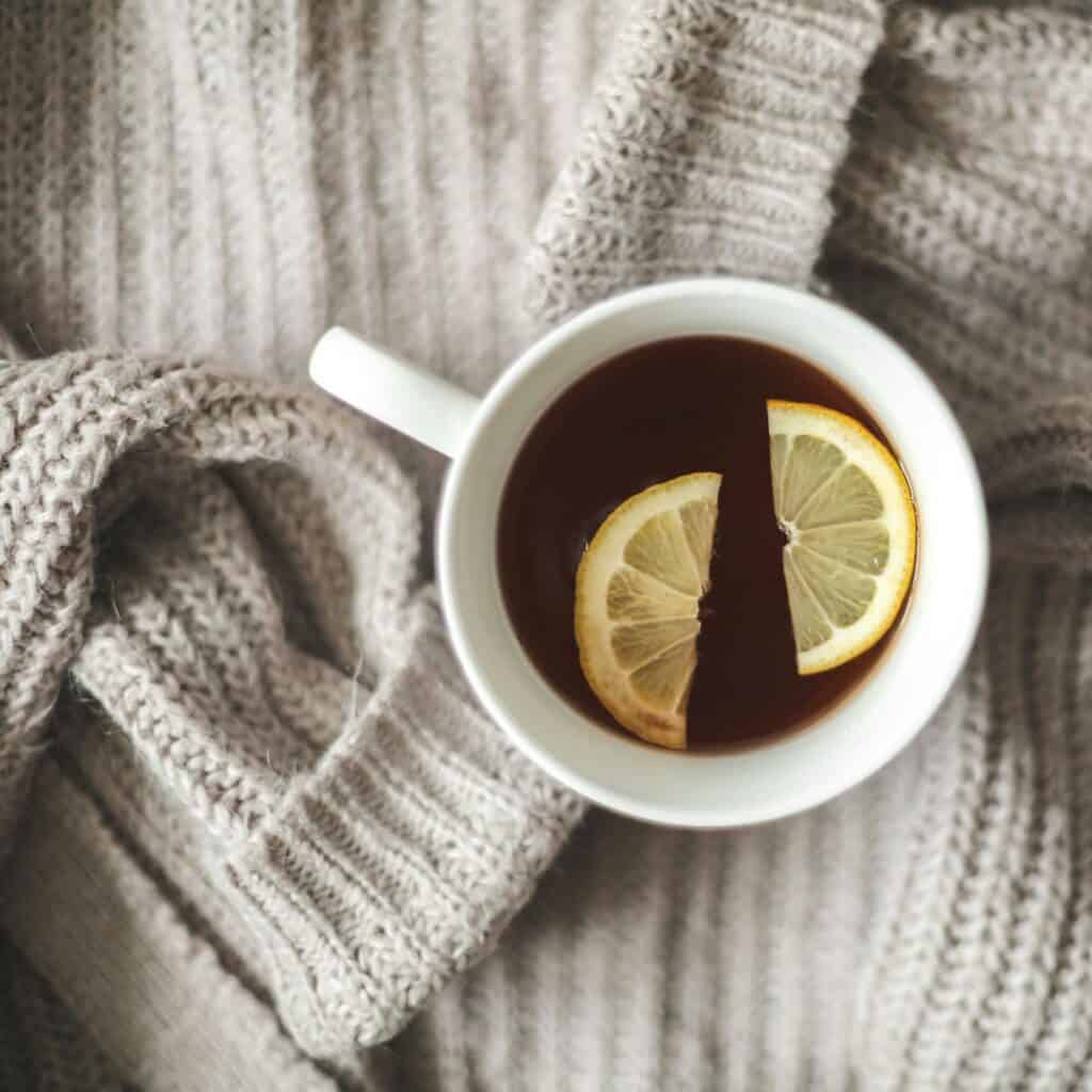A cup of tea with two lemon slices floating on top rests on a grey knit sweater.