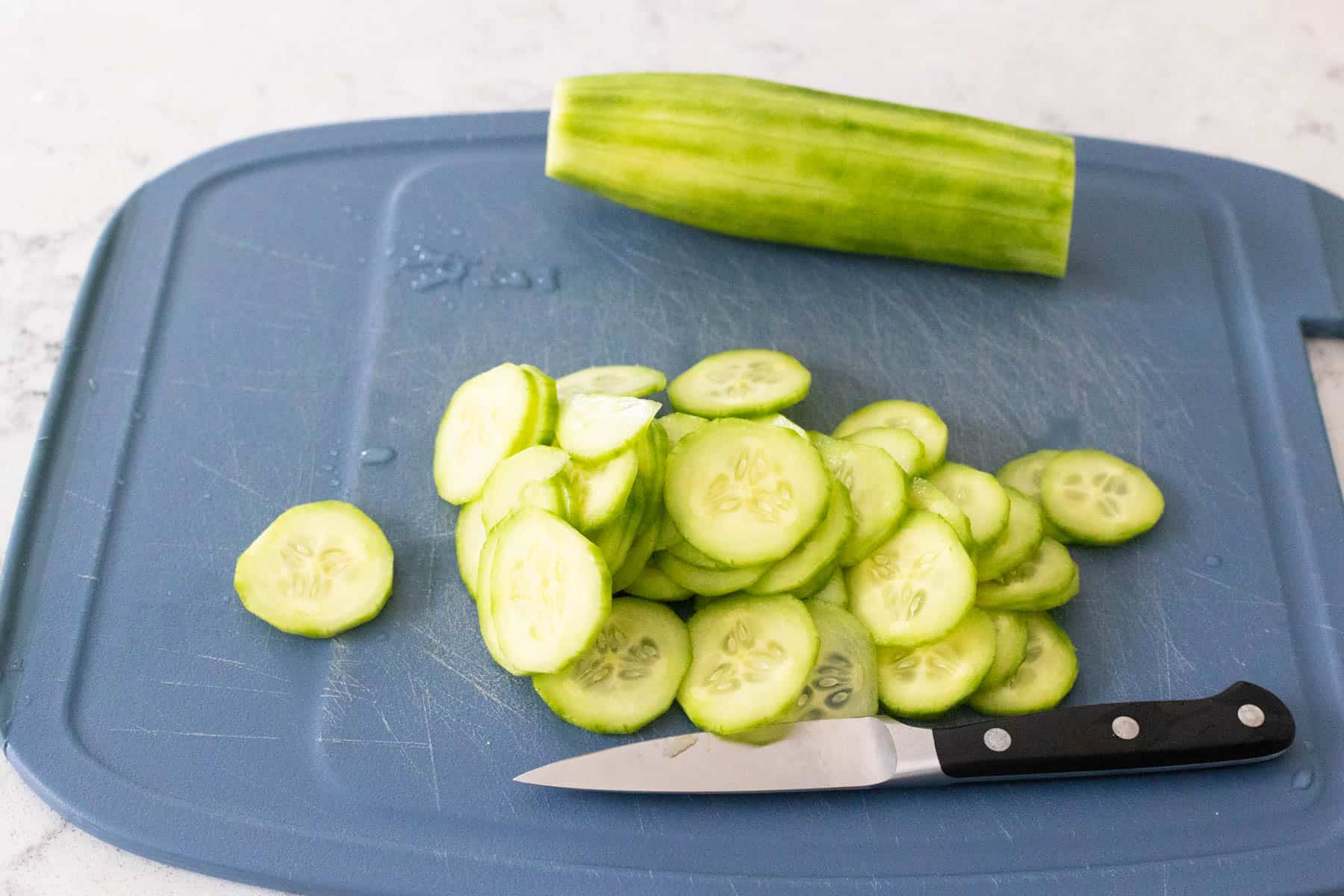 Shows how to prep the cucumbers by slicing them into thin rounds on a cutting board for zesty cucumber salad.