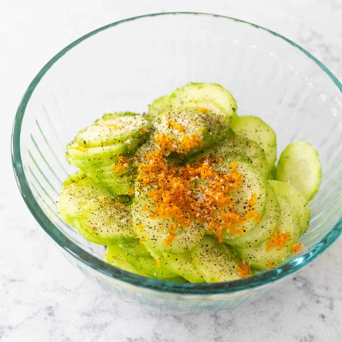 Sliced cucumbers in a glass bowl topped with salt, black pepper, and fresh orange zest for zesty cucumber salad.