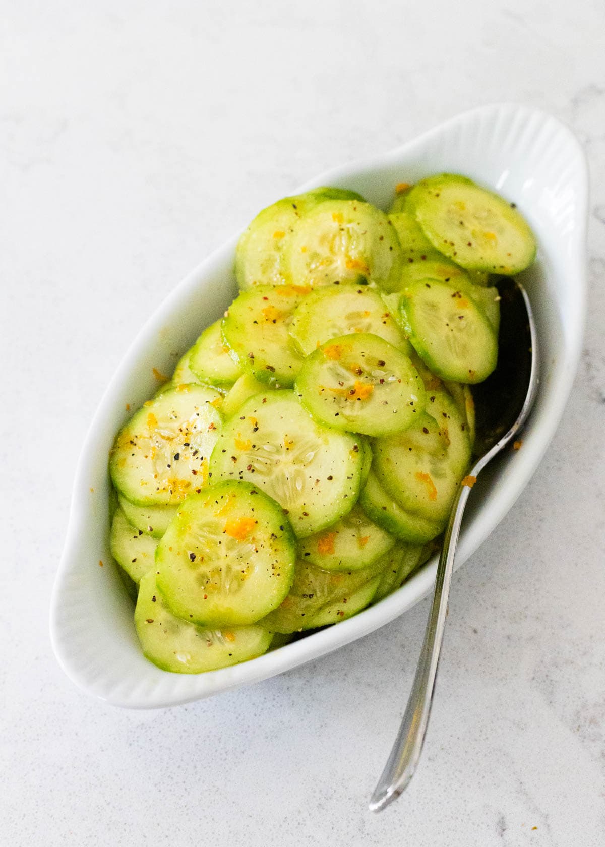 A white serving dish is filled with thinly sliced cucumber in a orange honey dressing.