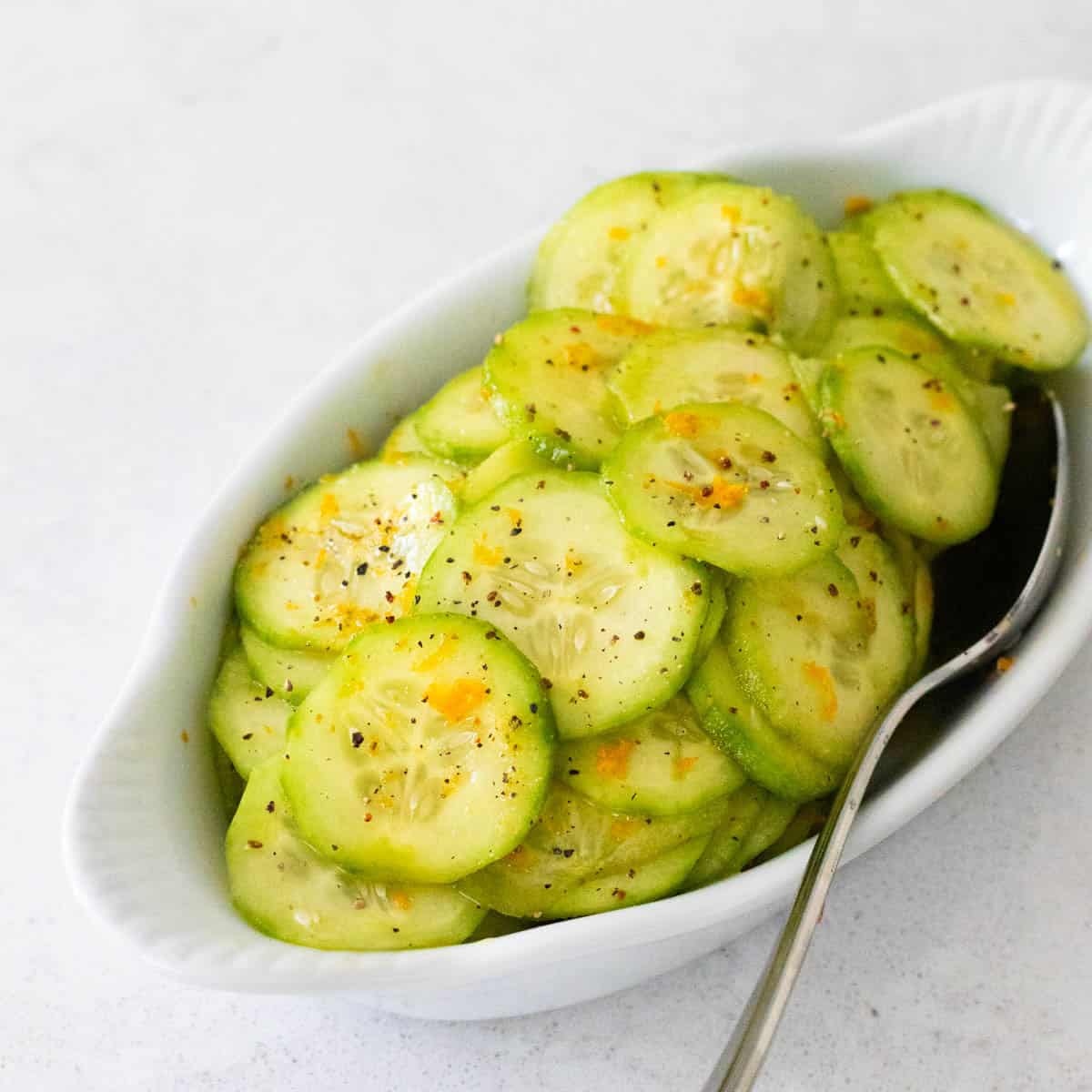 A white serving dish is filled with thinly sliced cucumber in a orange honey dressing.