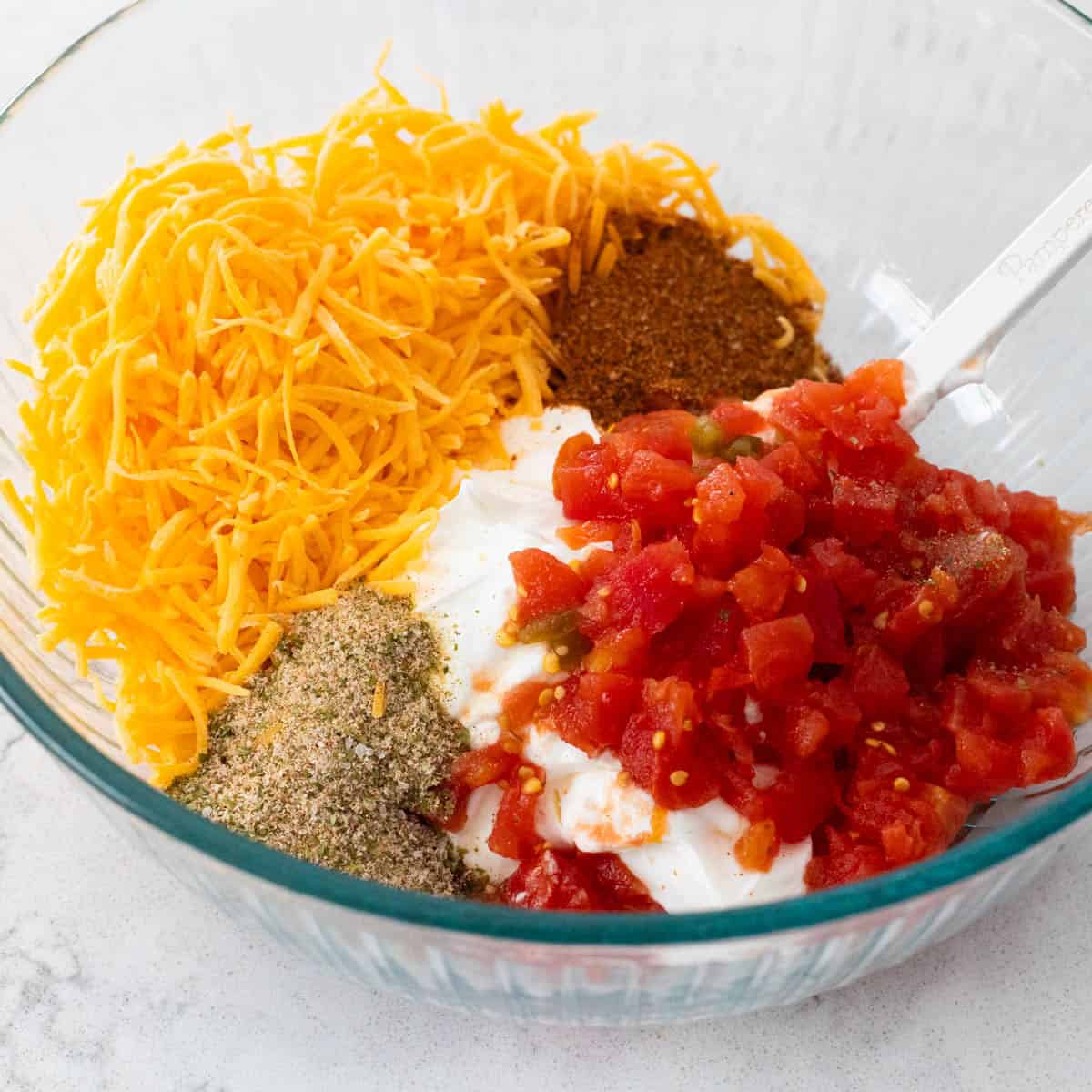 The cheese, tomatoes, spices, and sour cream are in the mixing bowl ready to be stirred together.