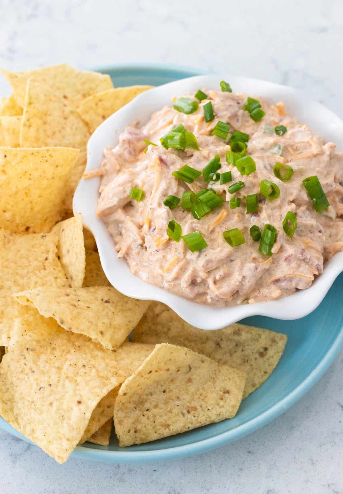 A white ruffled bowl is filled with the creamy boat dip with green onions sprinkled over the top. Tortilla chips are served for dunking.