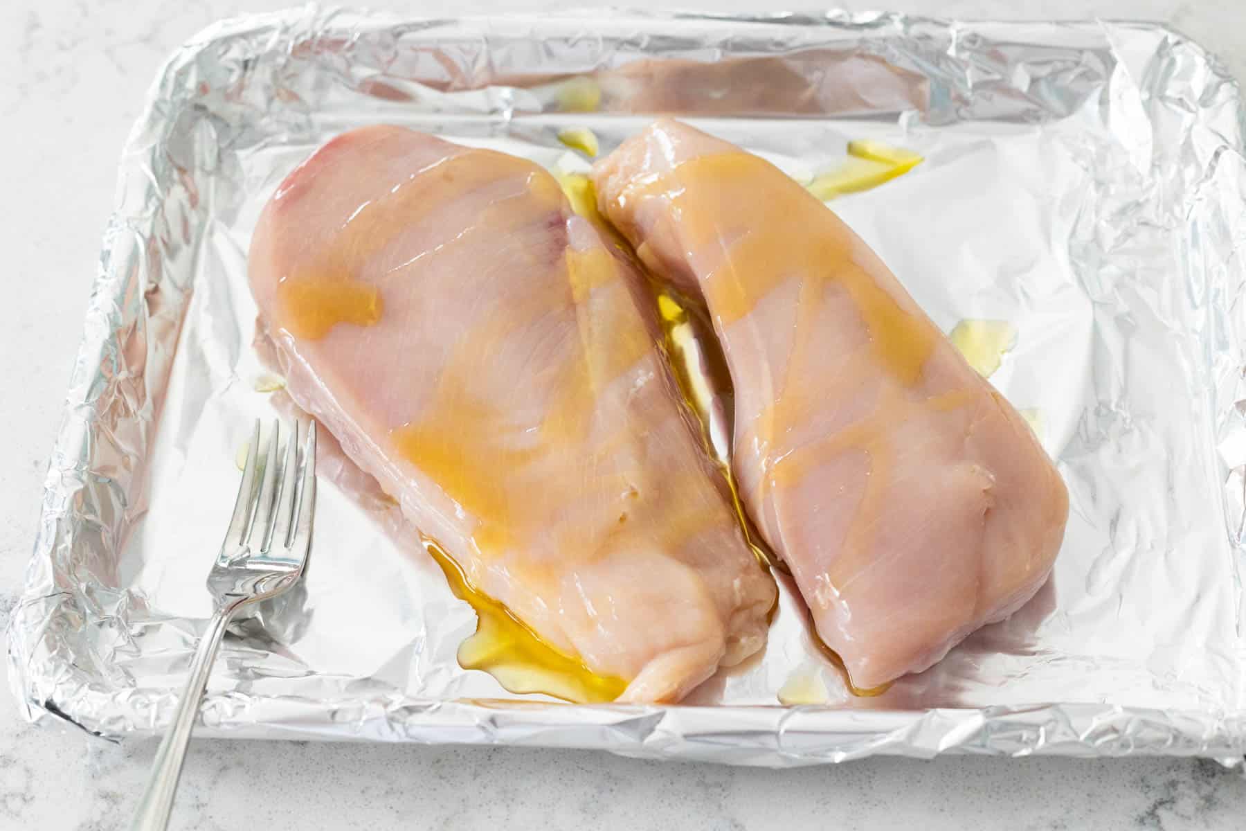 The olive oil has been drizzled over the raw chicken on a baking sheet.