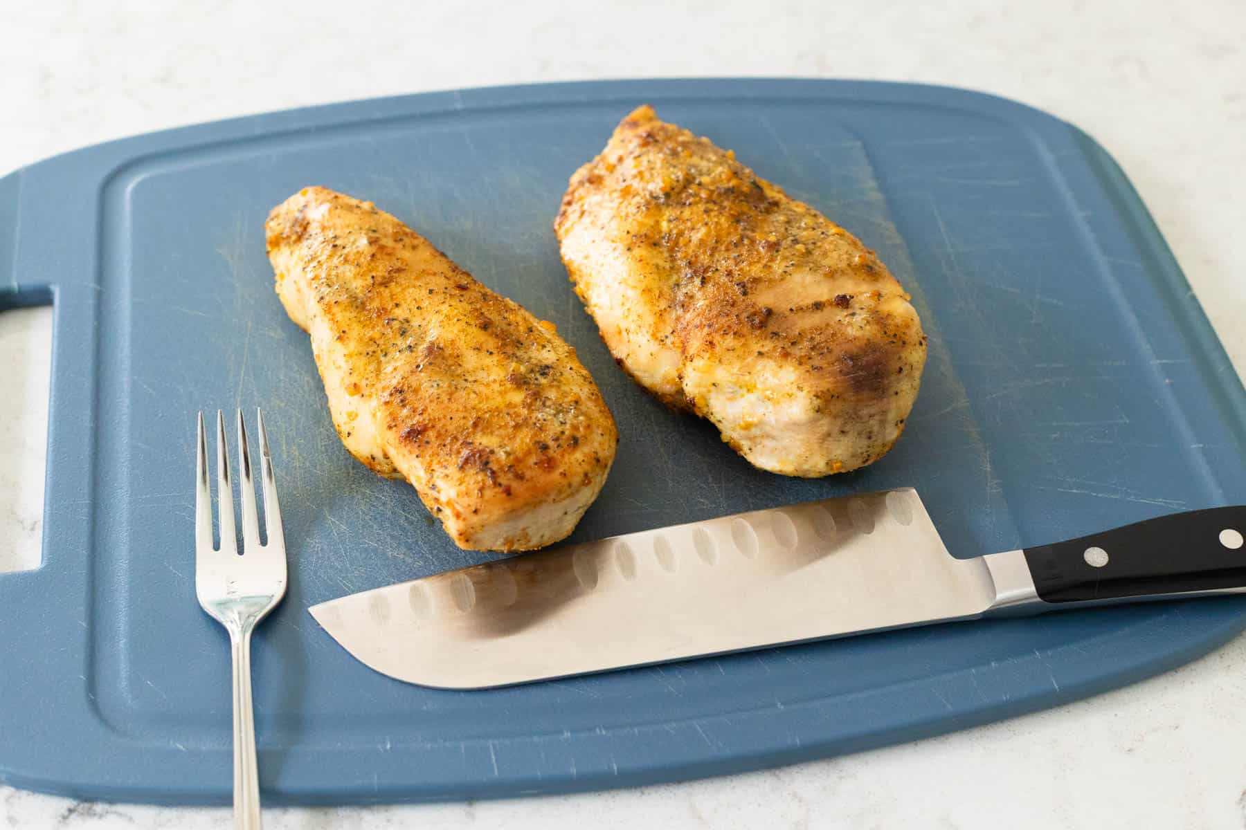 The chicken rests on a cutting board. There is a large knife and a fork waiting to do the slicing.