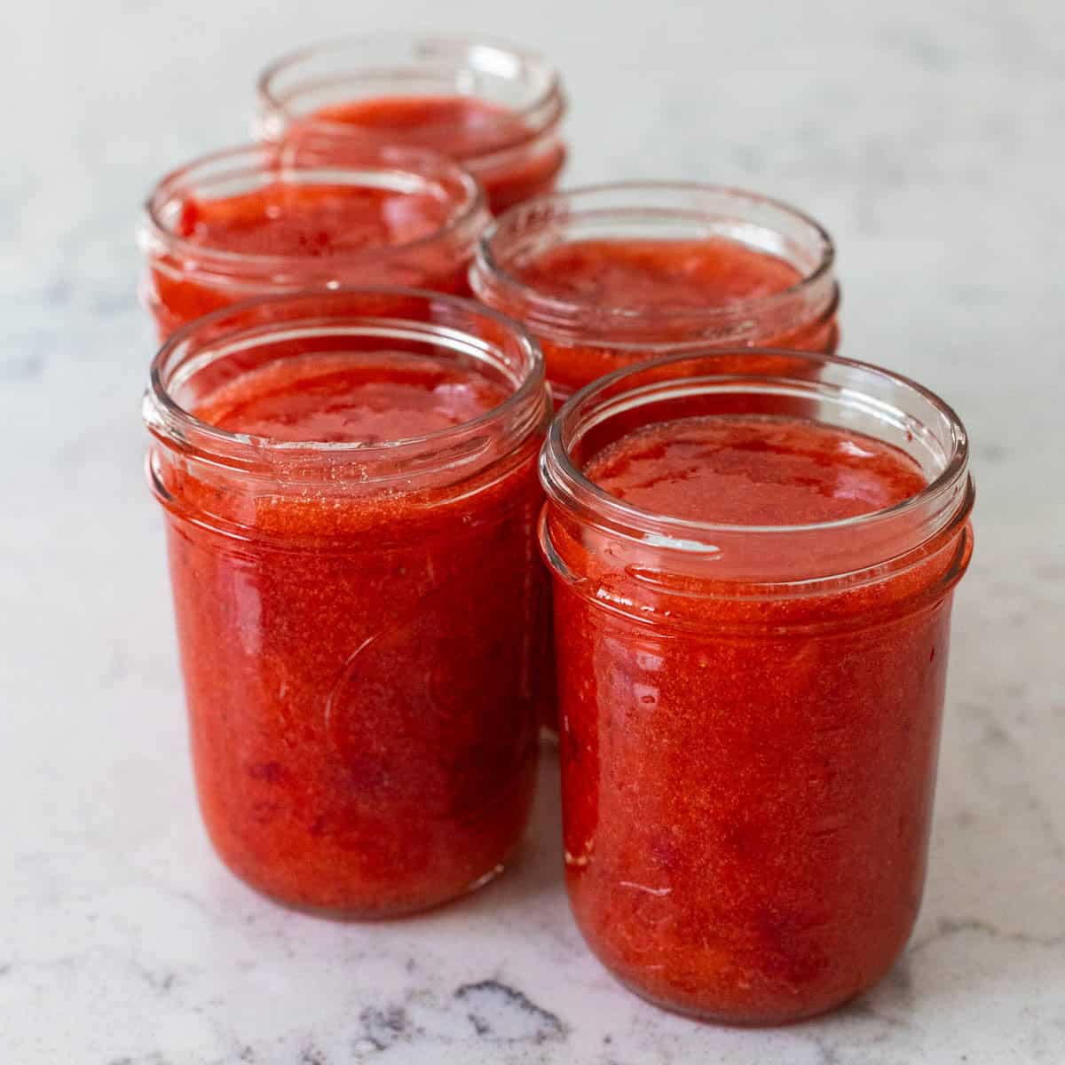 5 jars of strawberry jam are setting up on the kitchen counter.
