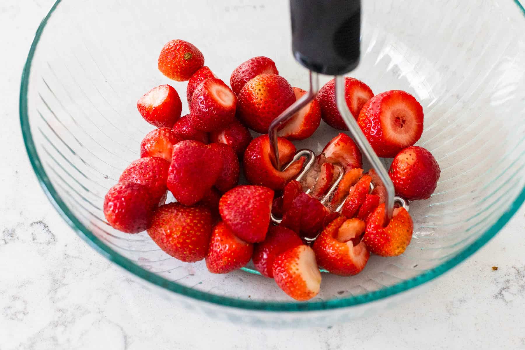 A potato masher is crushing the strawberries in a large mixing bowl.