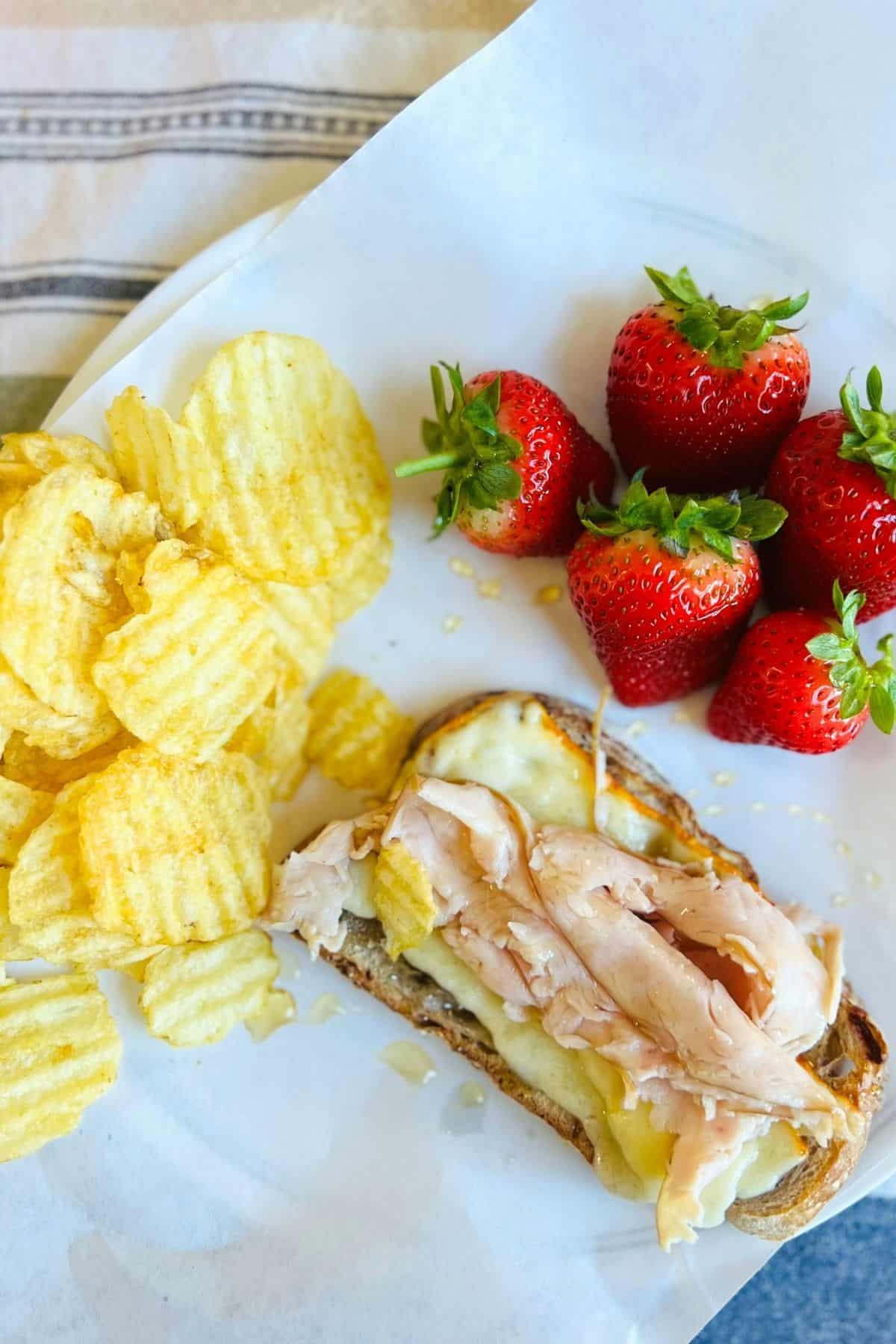 An open face sandwich sits on a piece of parchment paper with potato chips and fresh strawberries on the side.
