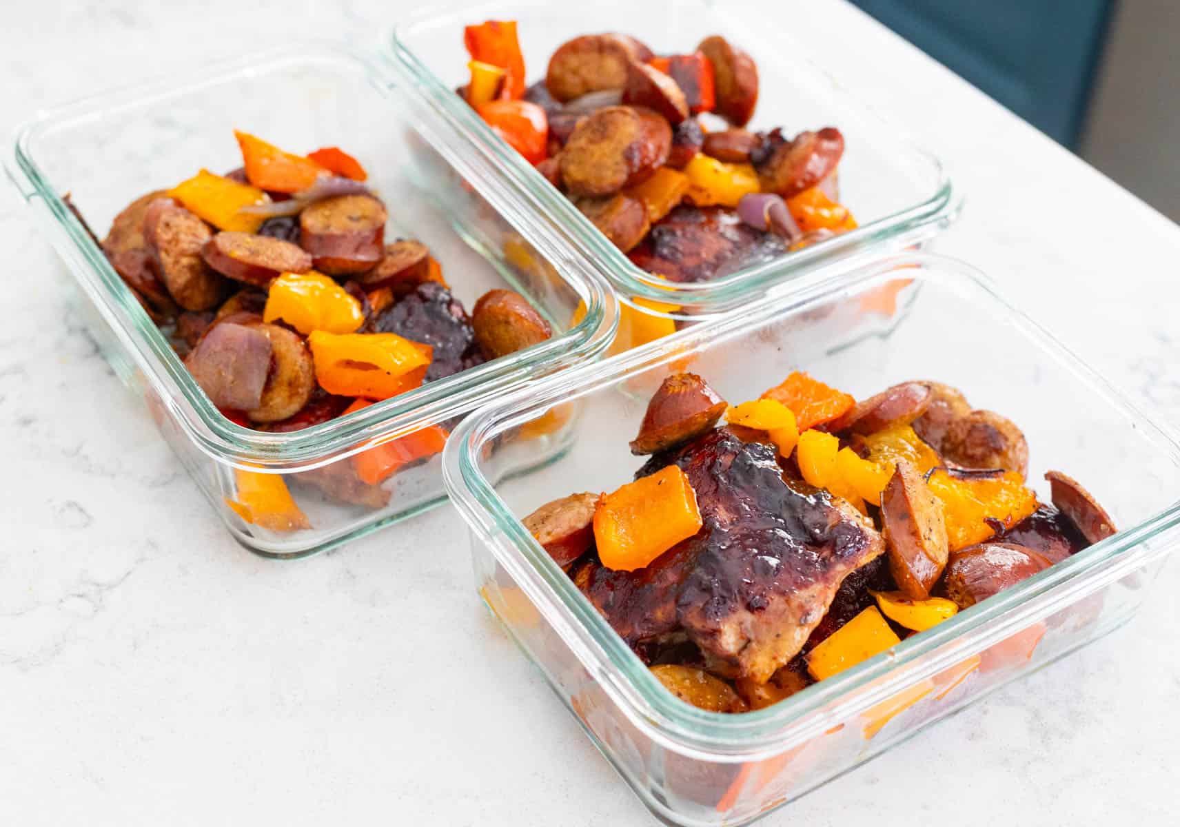 The baked barbecue chicken thighs and sausage and peppers have been portioned out into meal prep containers. 