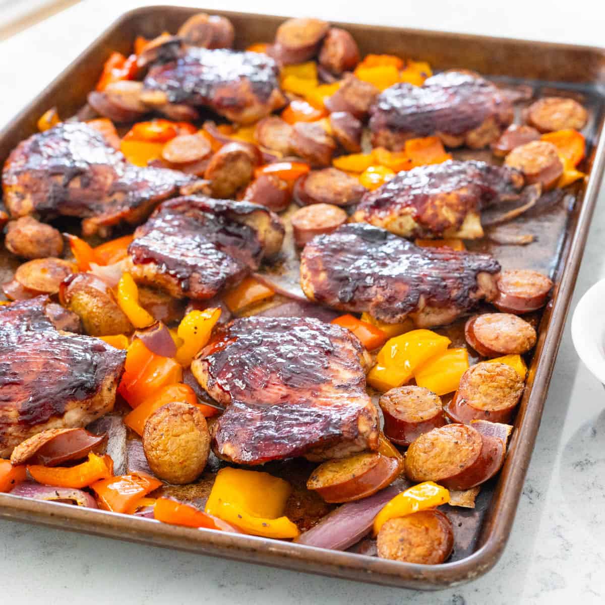 The sheet pan of baked barbecue chicken thighs, sausages, and peppers is resting on the counter. 