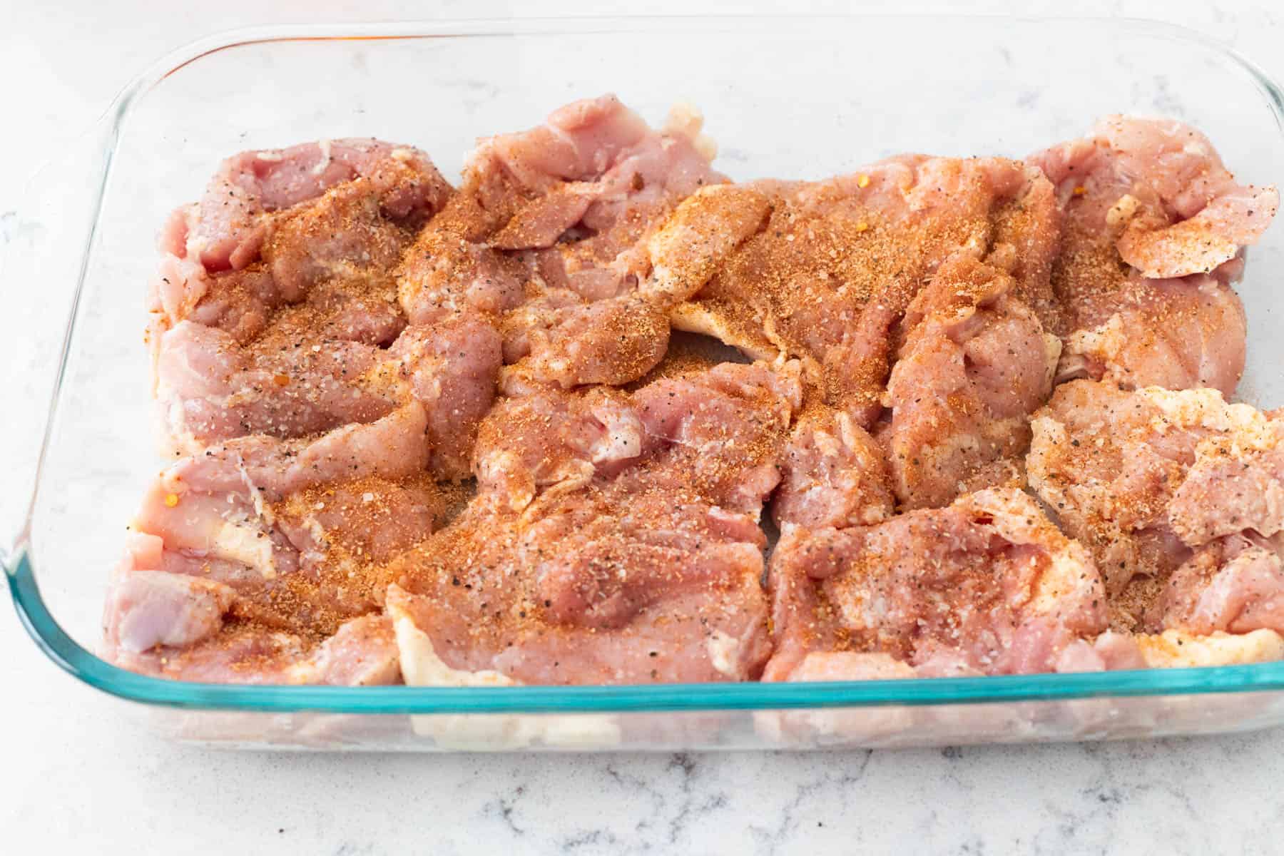 Chicken thighs are nestled in a 9x13 baking dish with seasoning on top. 