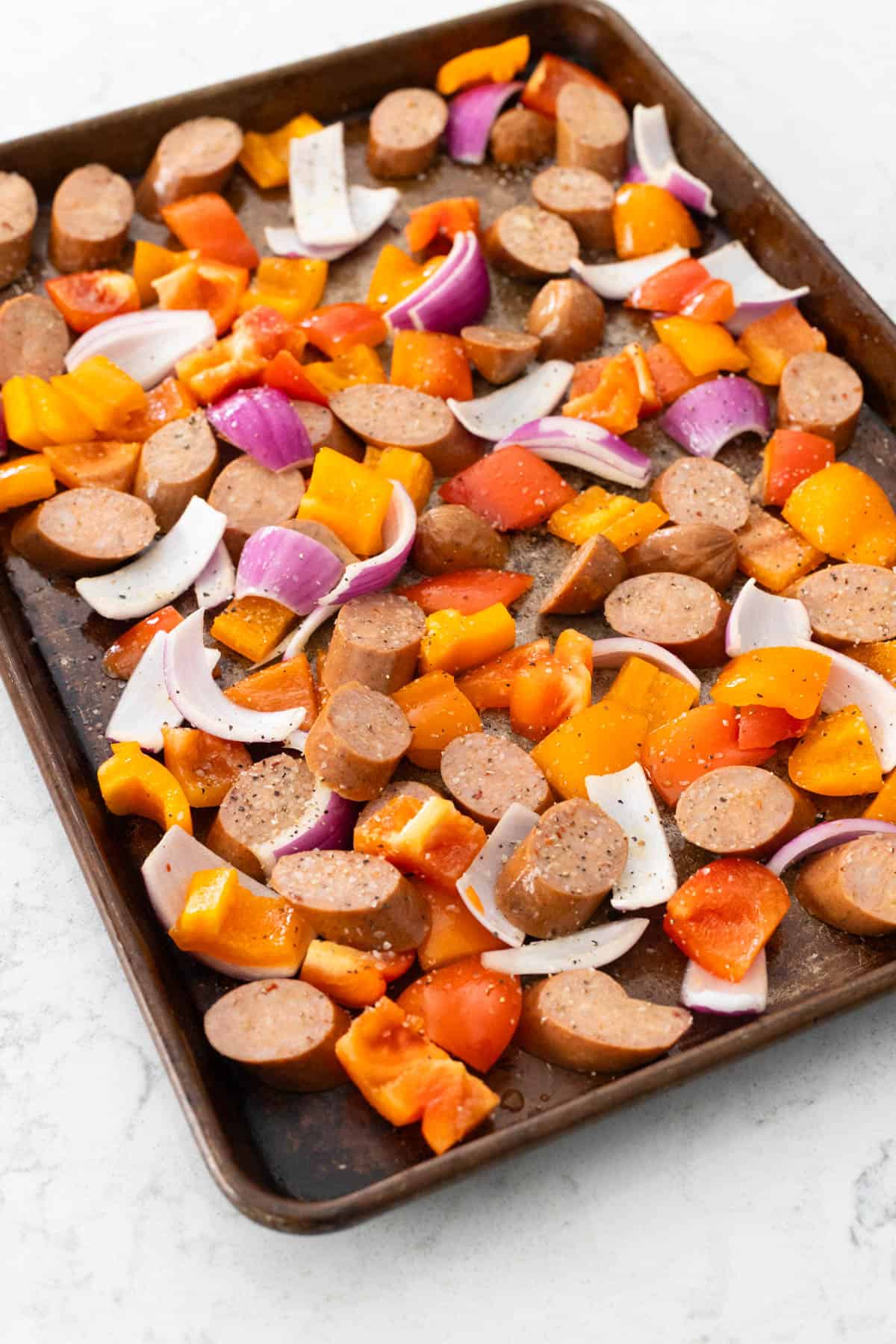 The baking sheet is filled with sliced sausages, peppers, and onions. 