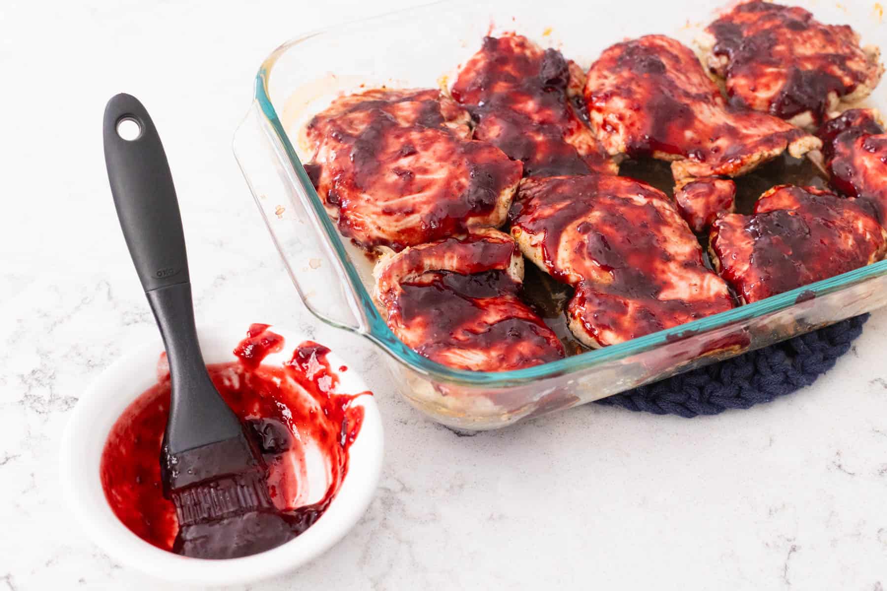 A cup of barbecue sauce with a pastry brush in it is sitting next to the baking dish filled with chicken thighs. 