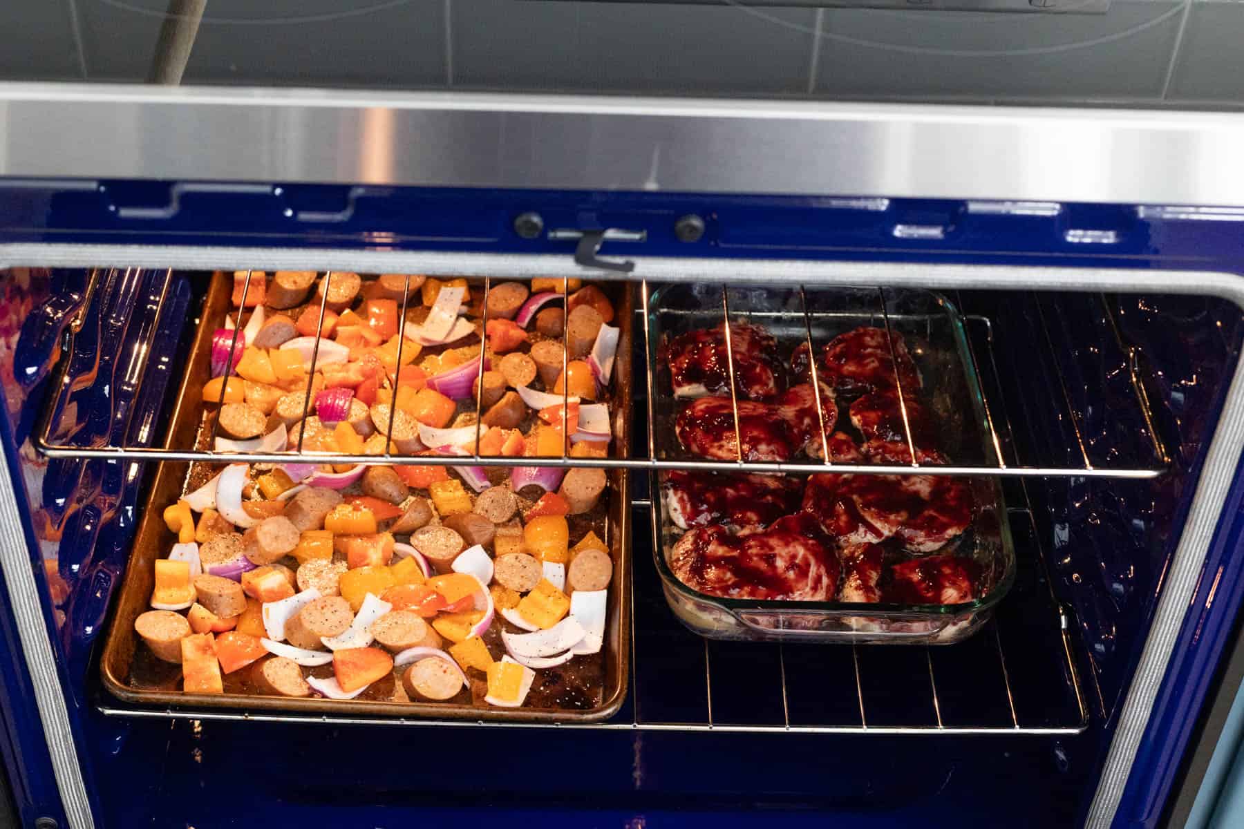 The sheet pan of sausages and peppers is on one side of the oven, and the baking dish filled with barbecue chicken thighs is on the other. They're baking at the same time. 