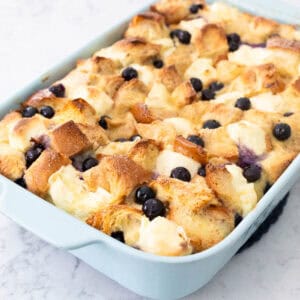 A blue baking dish is filled with a blueberry bread pudding with chunks of cream cheese dotted along the top.