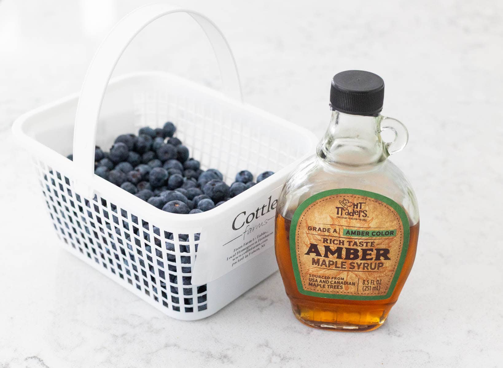 A farmer's basket of blueberries next to a jar of maple syrup.