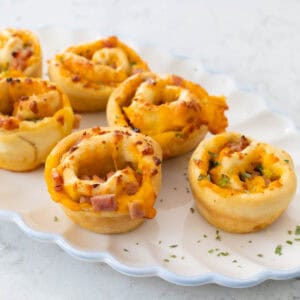 A white ruffled platter is filled with golden brown ham and cheese rolls.