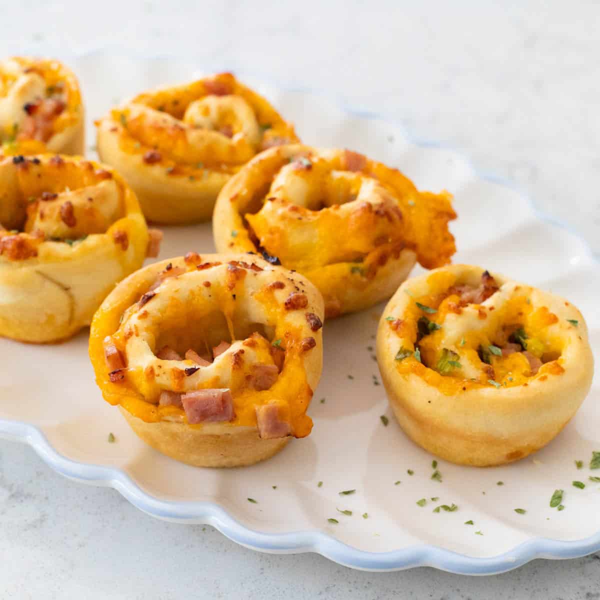 A white ruffled platter is filled with golden brown ham and cheese rolls.