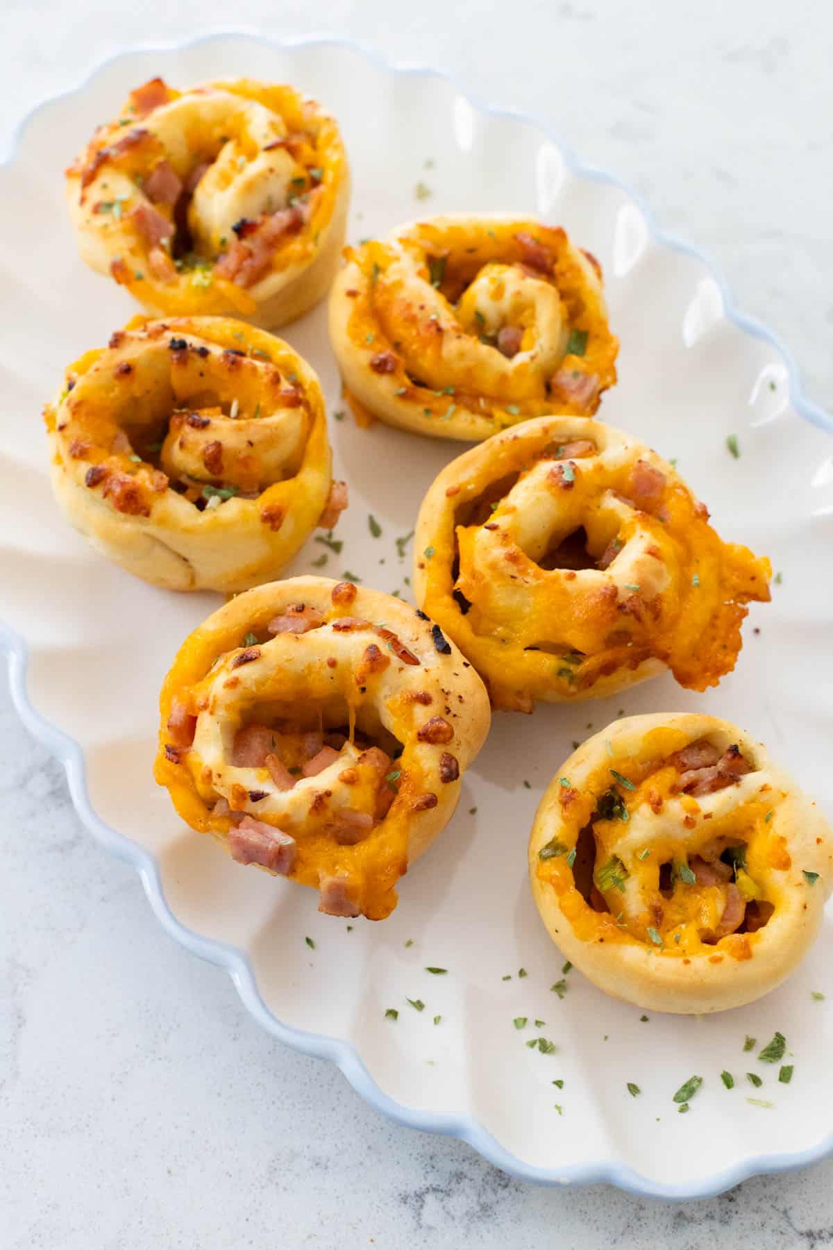 A white ruffled platter is filled with golden brown ham and cheese rolls.