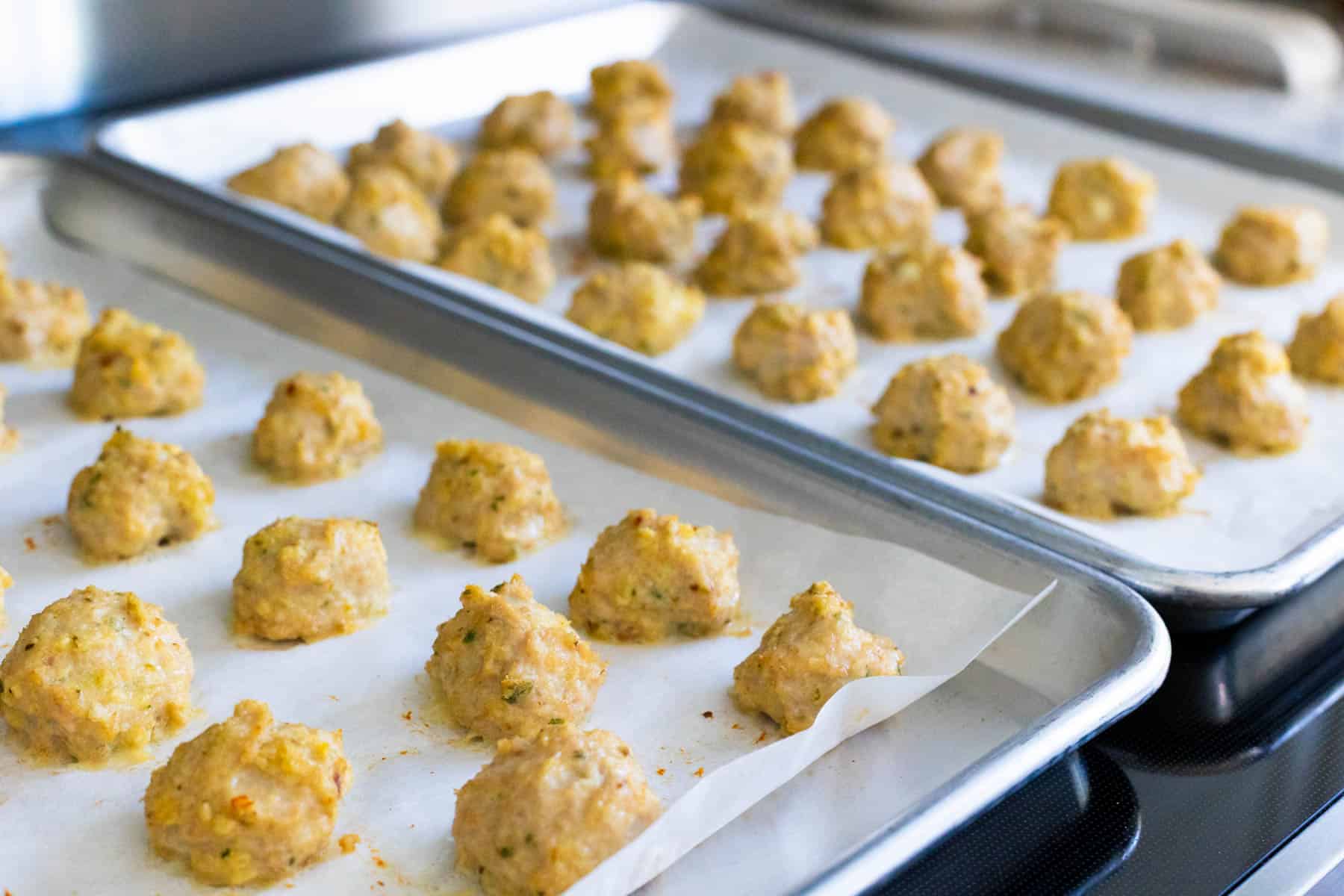 Two rimmed sheet pans lined with parchment paper are sitting on the oven. They have rows of freshly baked chicken meatballs cooling.