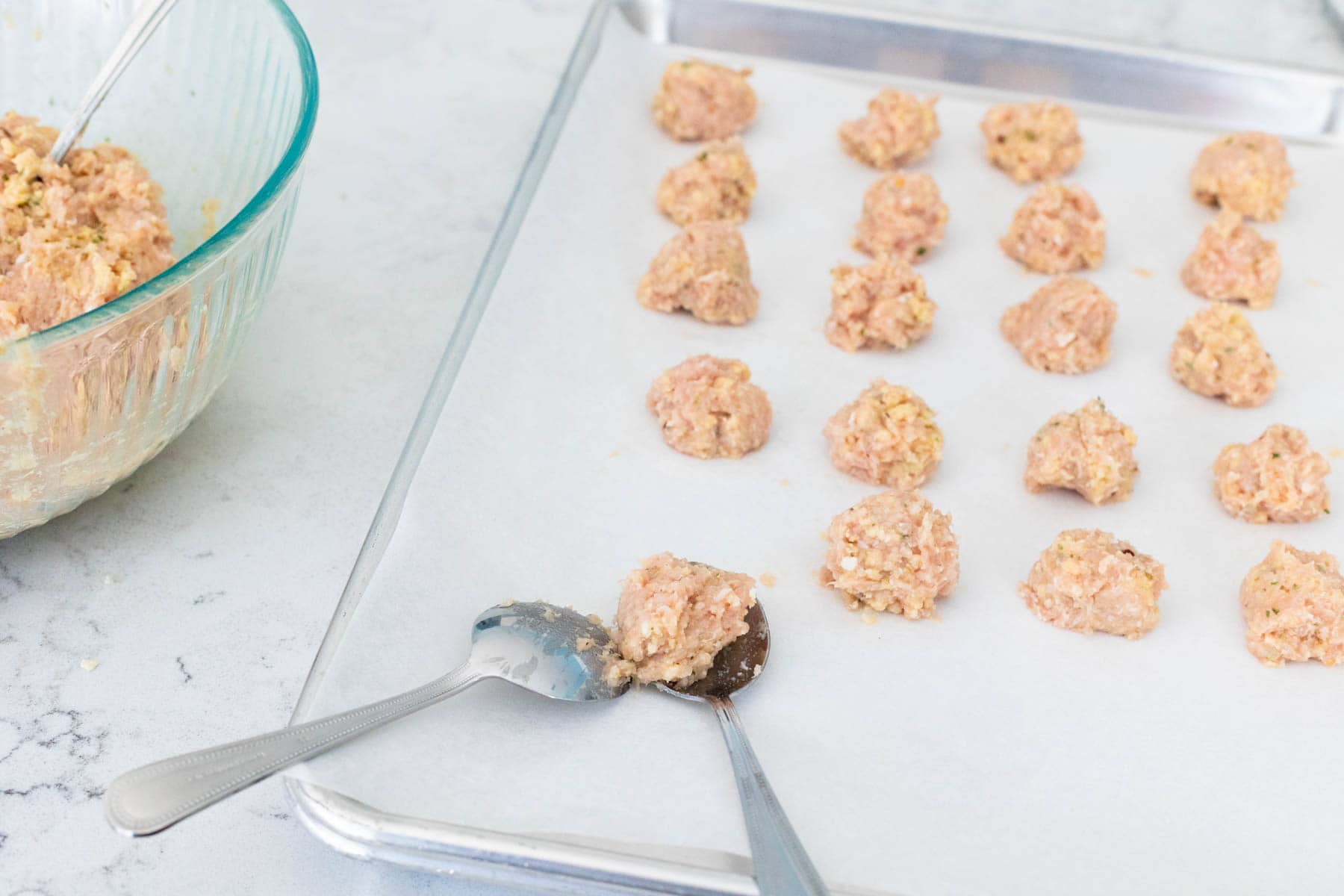 The meatball mix has been scooped by two spoons and formed on a sheet pan lined with parchment paper.