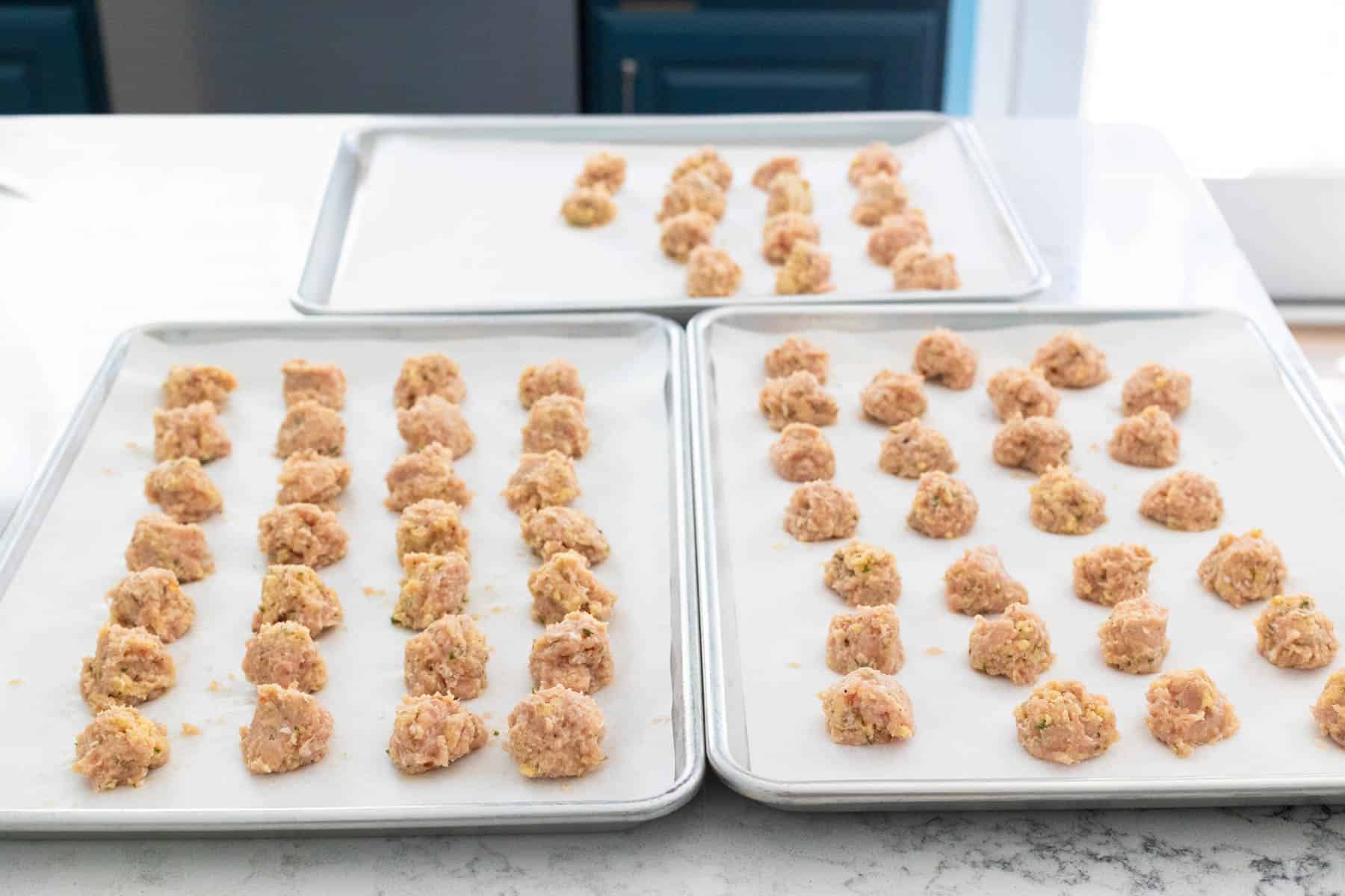 There are three baking pans lined with parchment filled with raw formed meatballs, on the kitchen counter.