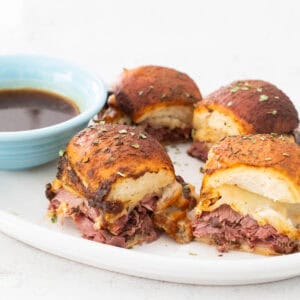 Freshly baked French dip sliders are served on a white platter with a blue cup of au jus for dipping.
